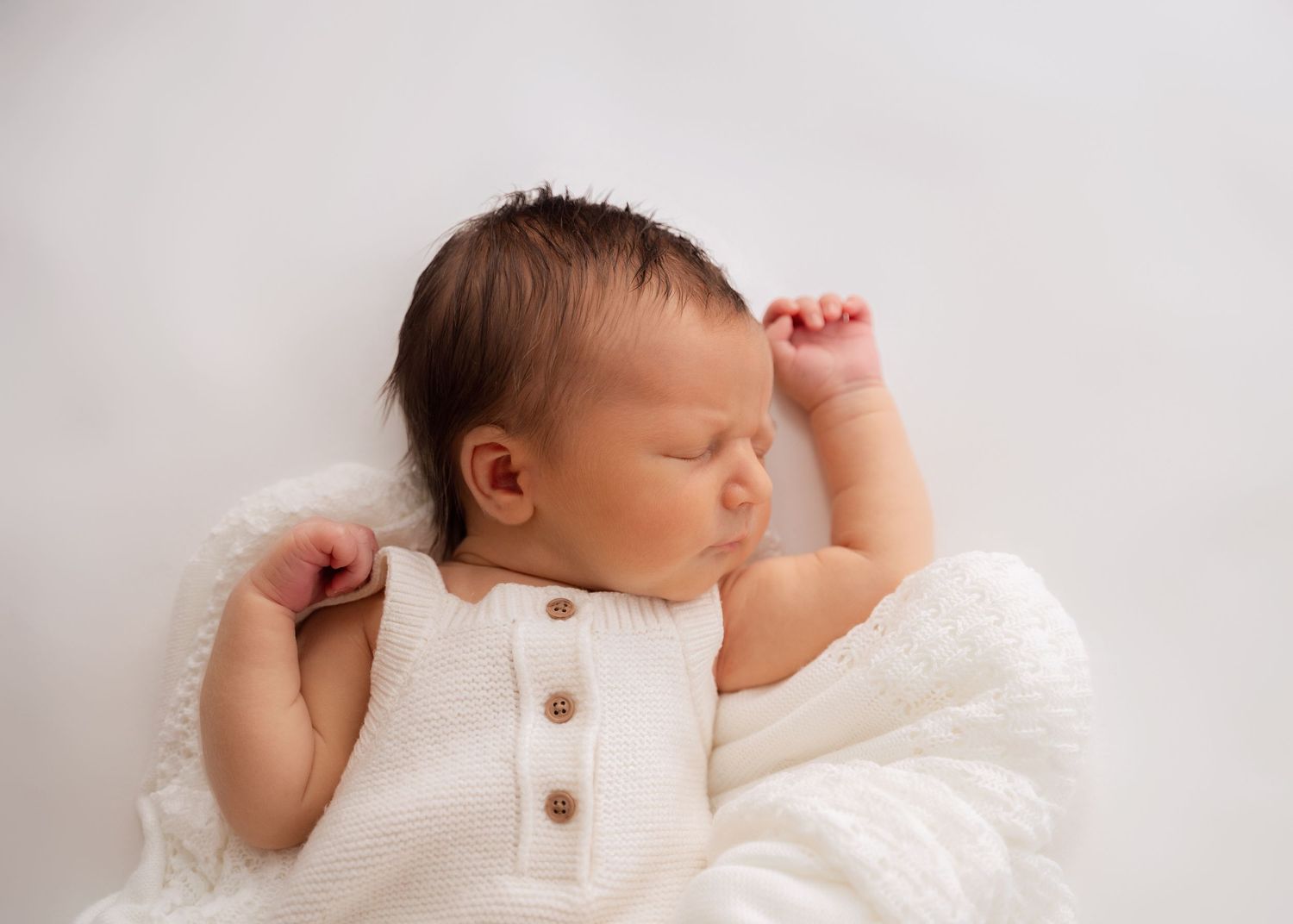Newborn baby lying on a white blanket with eyes closed and arms raised.