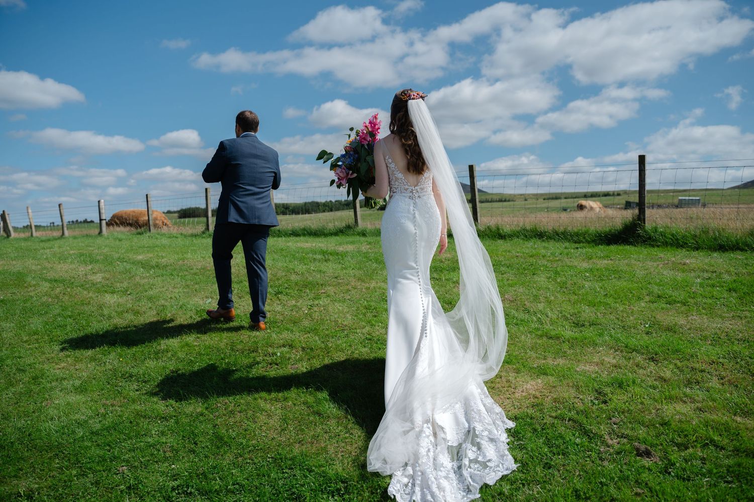 Bride and groom walking hand in hand across green field, veil flowing behind