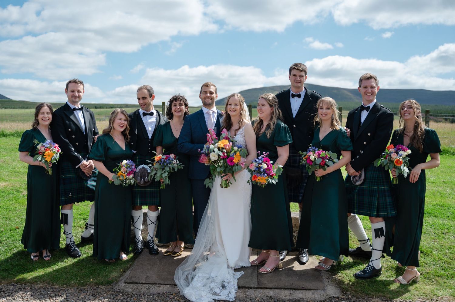 Bridal party smiling and seated outdoors with the bride and groom in the centre, holding colourful bouquets