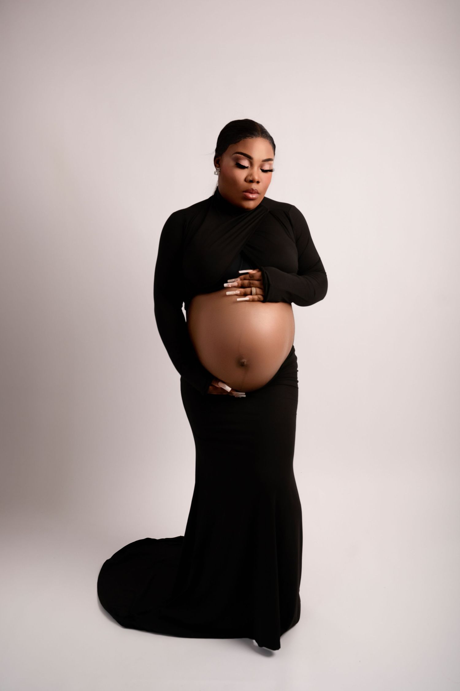 Pregnant woman in a long black gown holding her belly, standing confidently against a light studio backdrop.