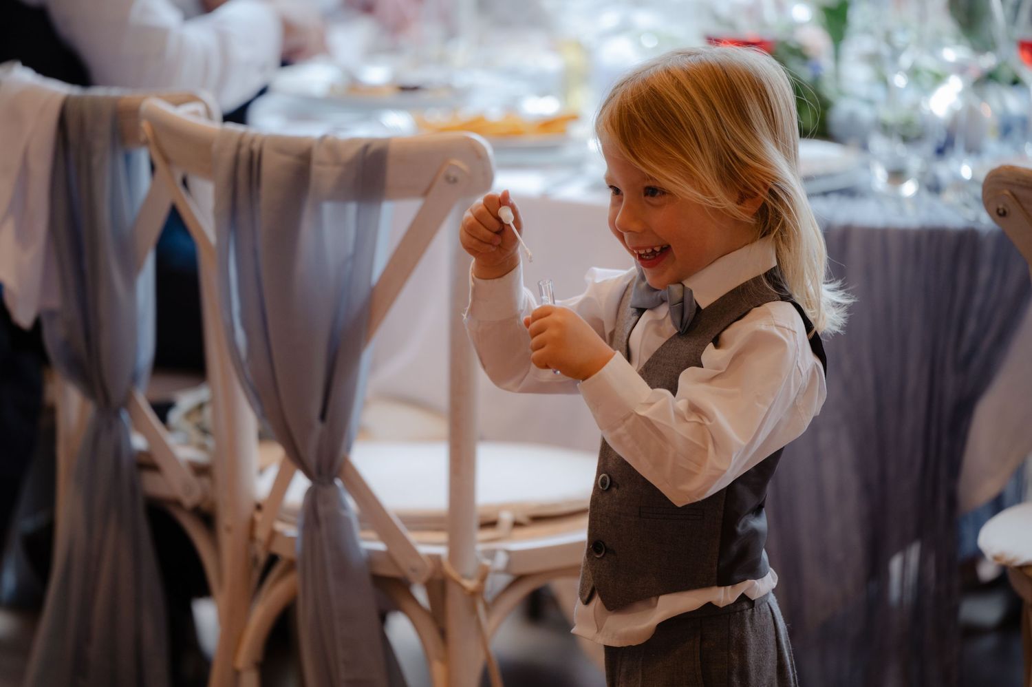 Young child playing with ribbon tied to a reception chair, dressed in a white shirt and waistcoat, with blurred tables in the background.