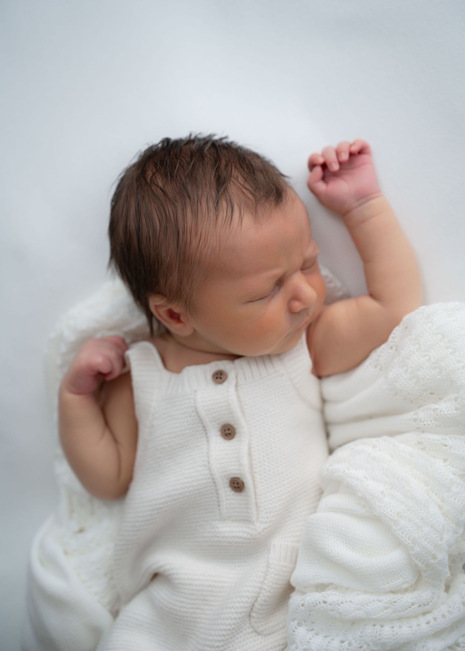 Newborn baby in a white knitted romper sleeping on a white backdrop.