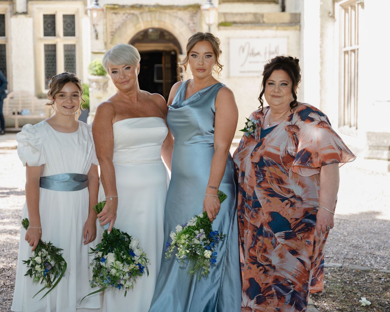Bride and bridesmaid holding their bouquets, smiling warmly at each other outside the venue, dressed in white and light blue gowns.