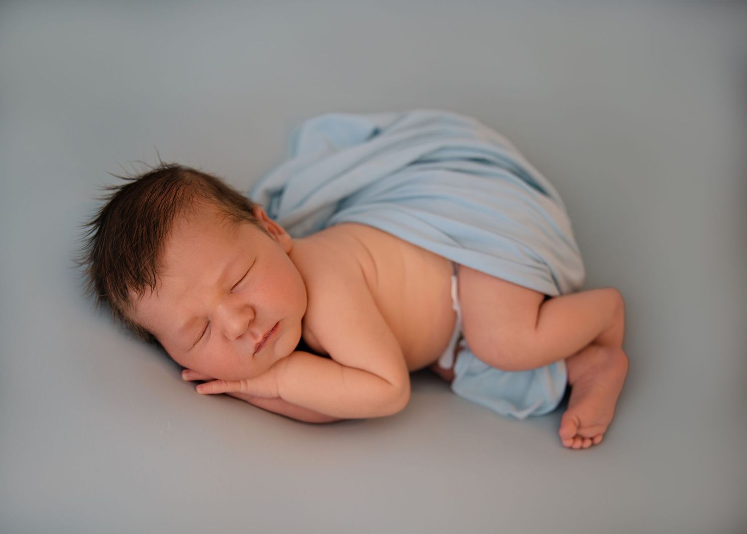 Newborn baby peacefully sleeping on a pale surface, covered partially with a light blue swaddle.