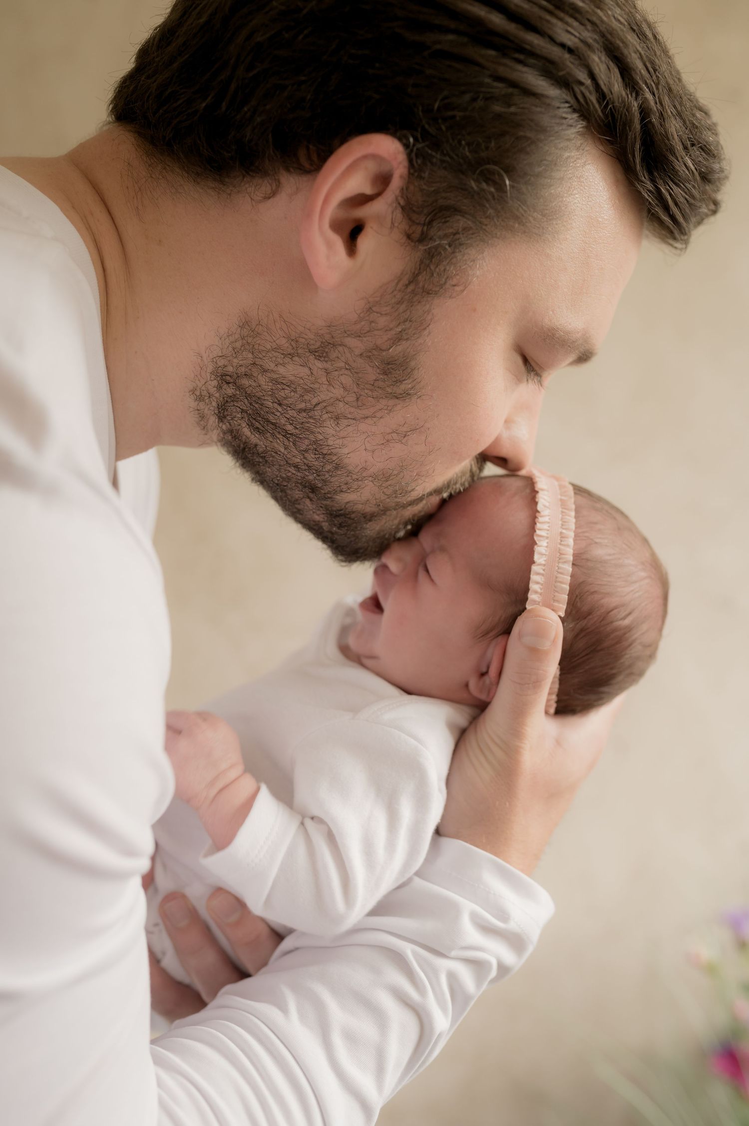 Father gently kissing his newborn baby’s forehead while holding them close in a tender moment, both dressed in soft neutral tones.