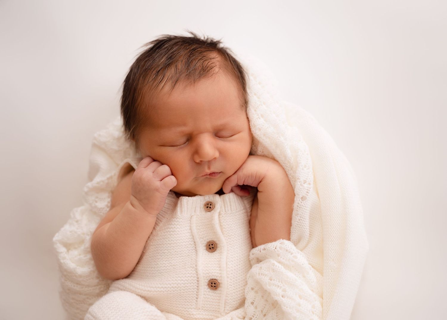 Newborn wearing a white outfit with hands near face.