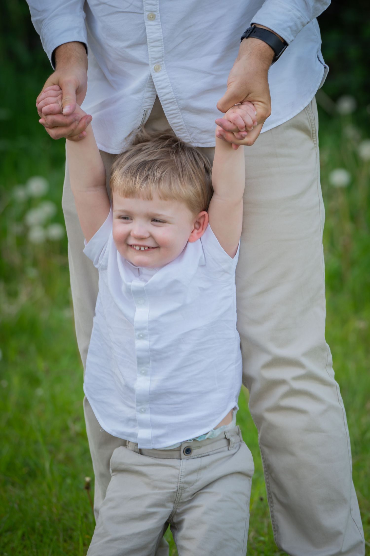 Little boy laughing as he is playfully lifted by both arms in a grassy outdoor setting, dressed in white shirt and beige trousers.