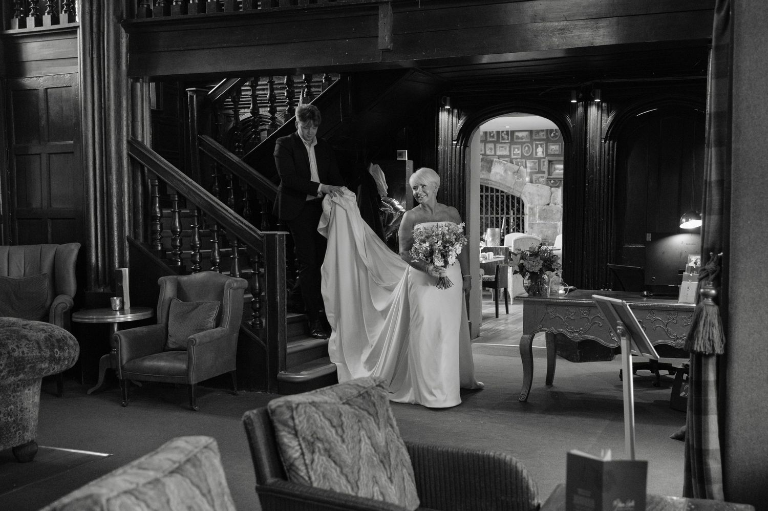 Black and white photo of the bride standing in an elegant, vintage-style hotel room, adjusting her dress before the ceremony.