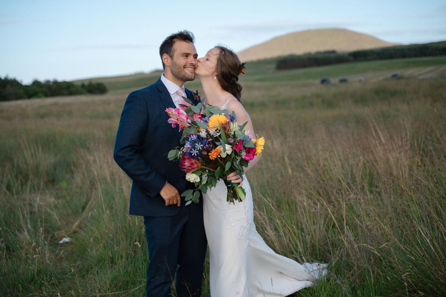 Newlyweds smiling and holding colourful bouquet in countryside setting