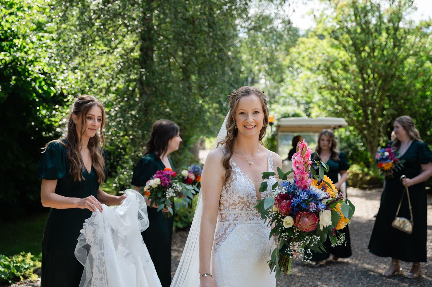 Bride smiling as she walks with her bridesmaids outdoors on a sunny day, holding colourful flowers