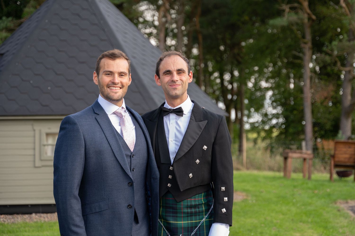 Groom standing with groomsman in a kilt, smiling outside near a wooden cabin