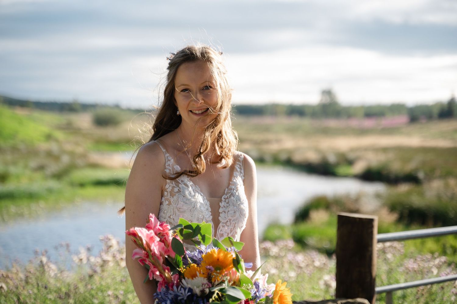 Bride smiling in golden sunlight near fence with wildflowers and water behind