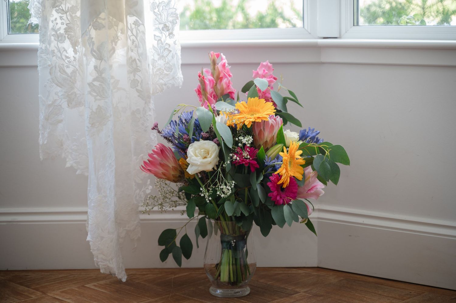 Large bridal bouquet placed in a glass vase near a bright window with sheer curtains