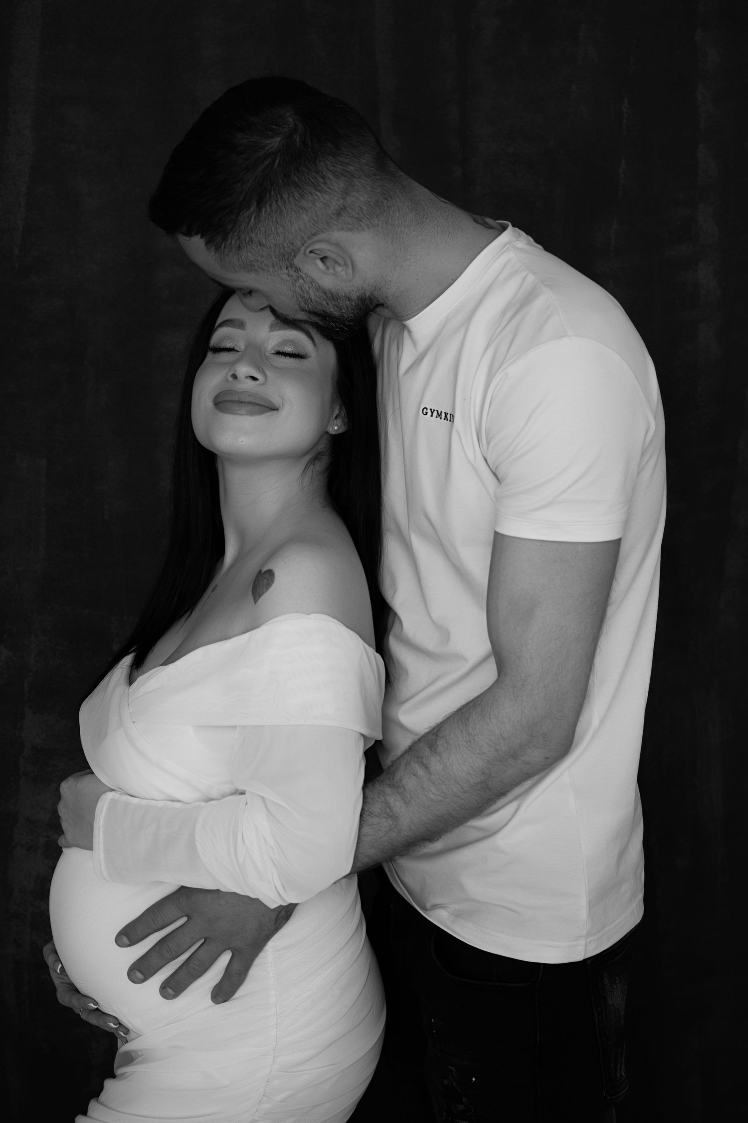 Intimate black and white maternity portrait of a couple. The man kisses the woman’s forehead while holding her belly.