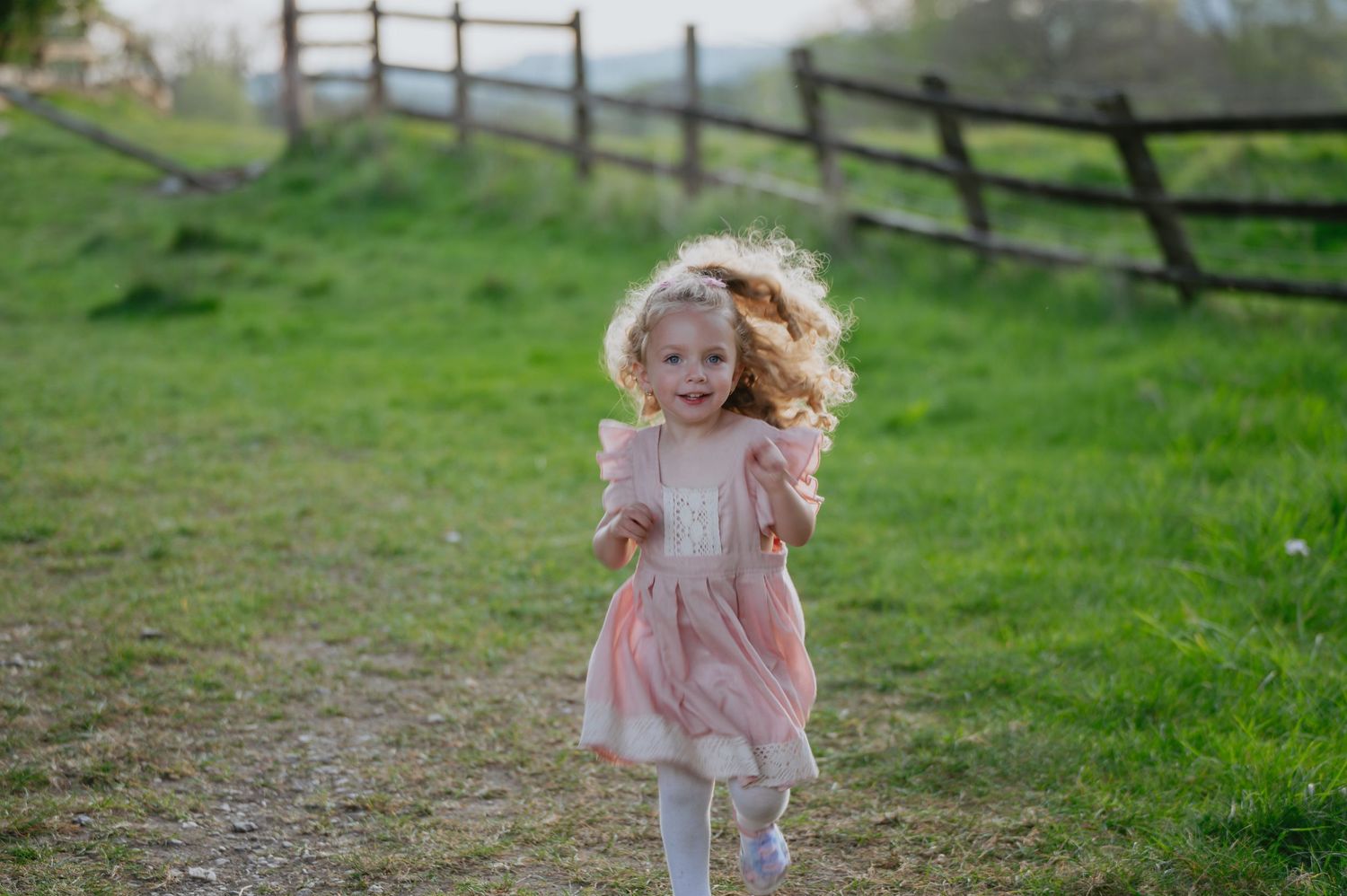 Little girl in a pink dress standing confidently in a field with a rustic wooden fence behind her, bathed in soft evening light.