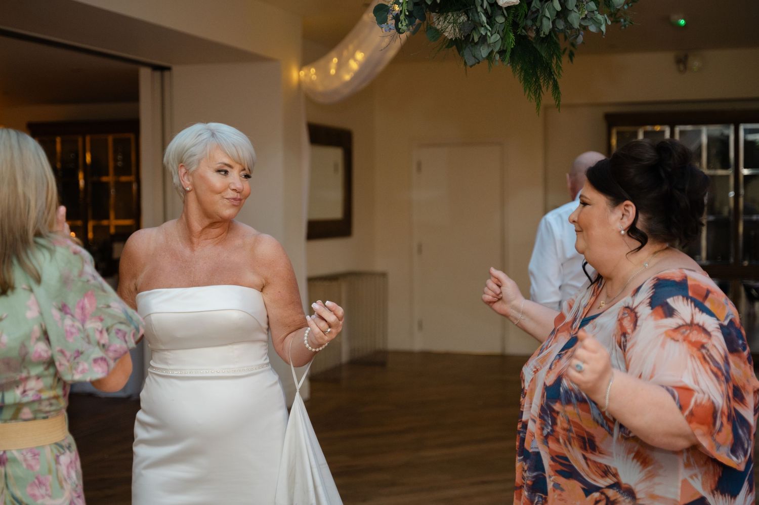 Bride in a strapless white dress chatting and laughing with wedding guests inside a warmly lit reception venue.