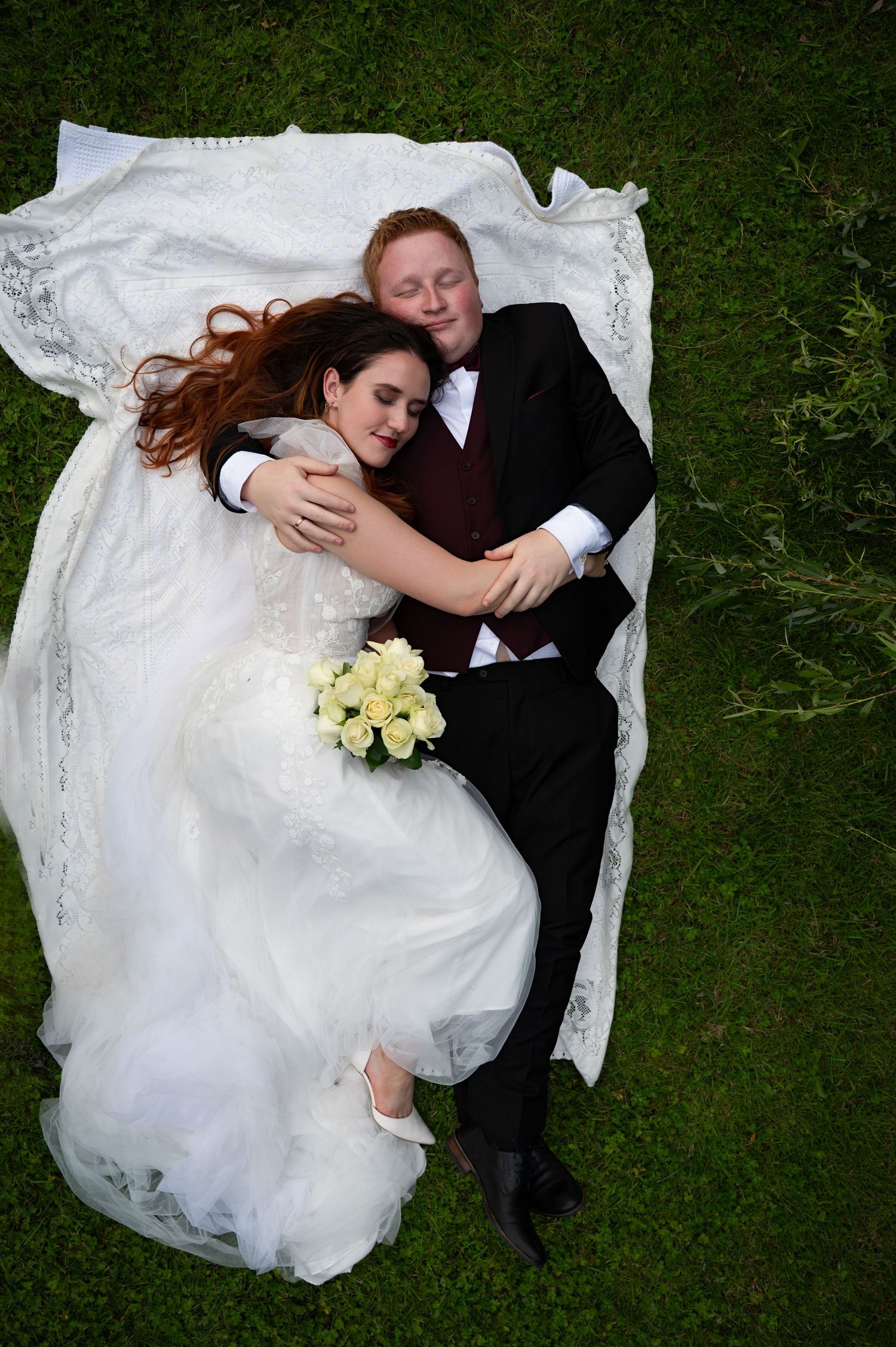 Bride and groom lying on a blanket in the grass, cuddled close and smiling