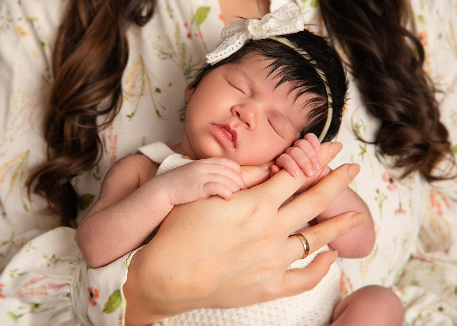 Newborn baby girl lying on a floral blanket, touching a parent’s hand, wearing a delicate headband with dark hair styled neatly.