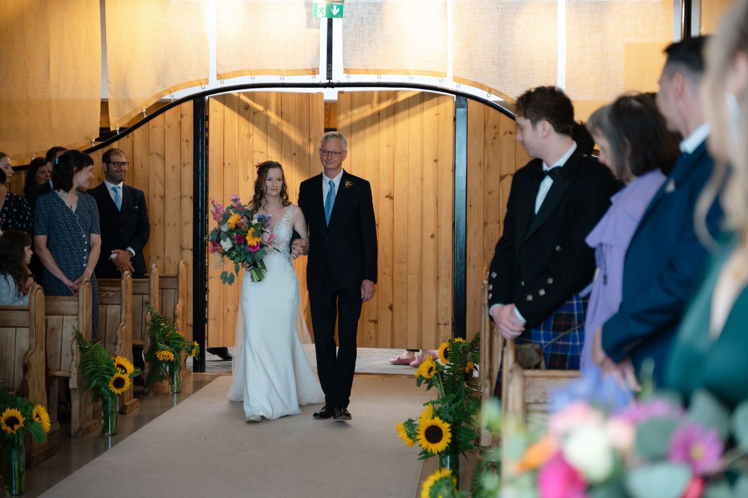 Bride walking down the aisle with her father, surrounded by standing guests and sunflower decorations