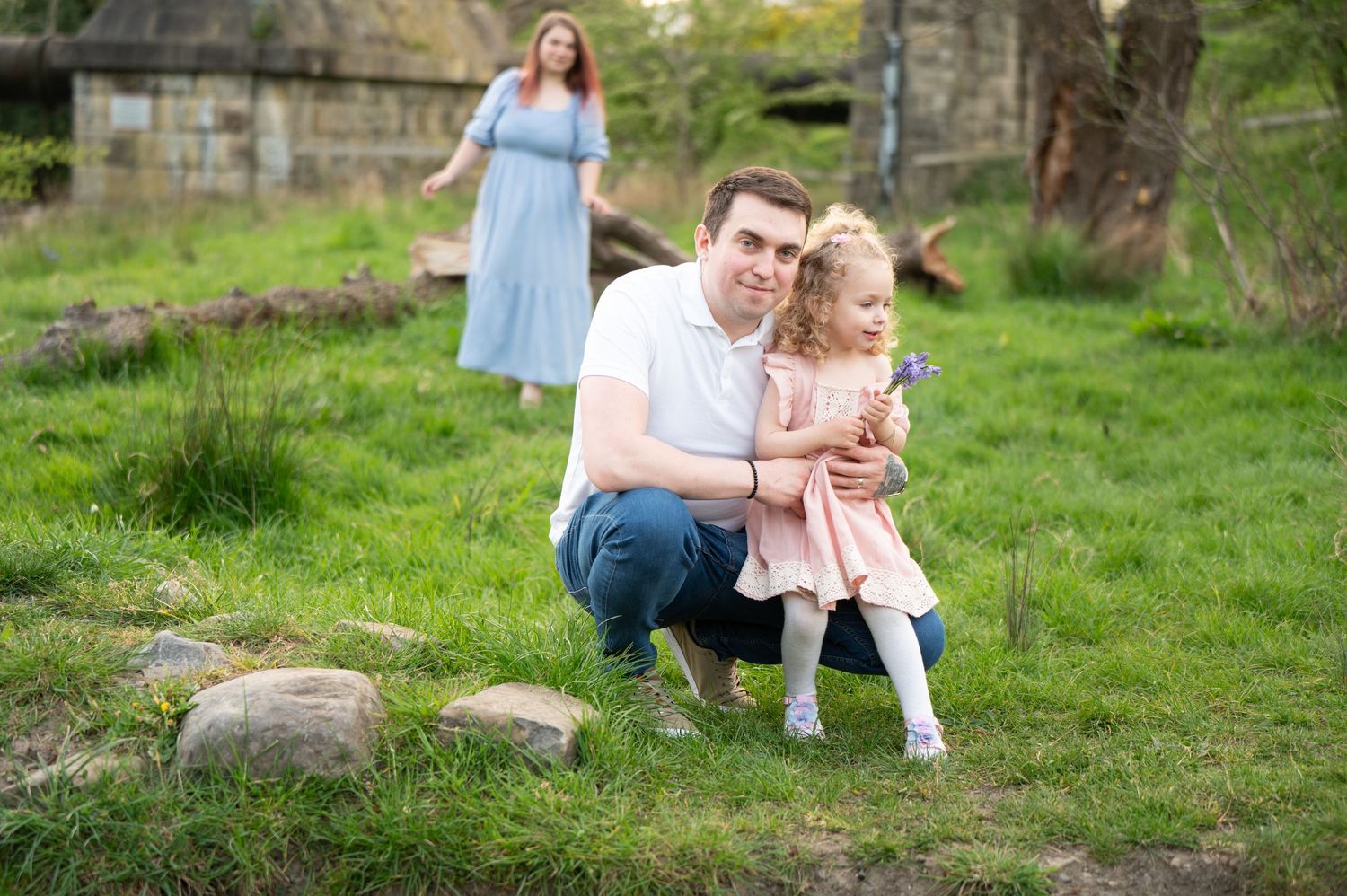 Father crouching in the grass holding his daughter on his knee, while mother walks in the background in a long blue dress.