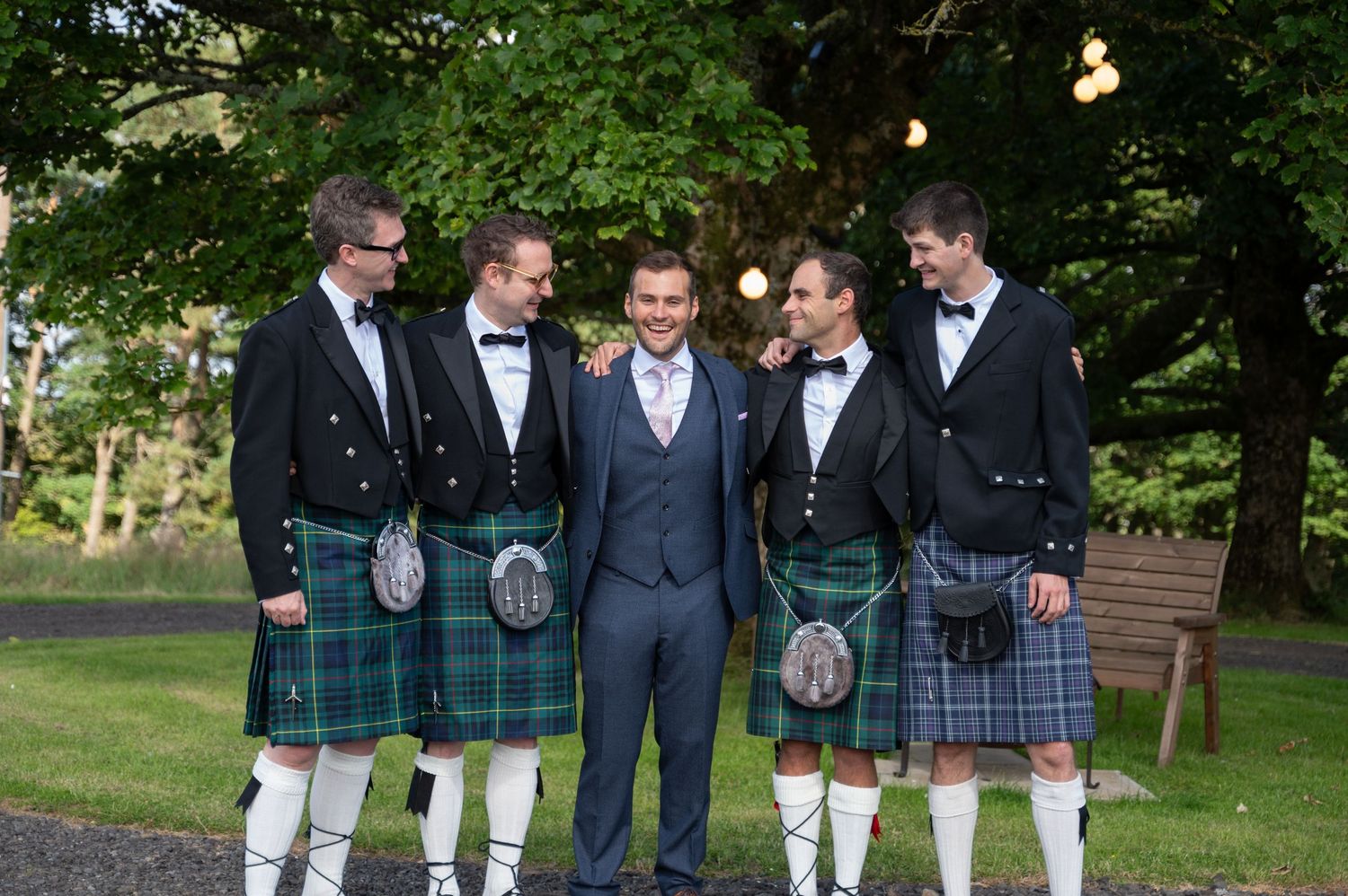 Groom laughing with groomsmen wearing kilts and suits, standing under trees in a garden