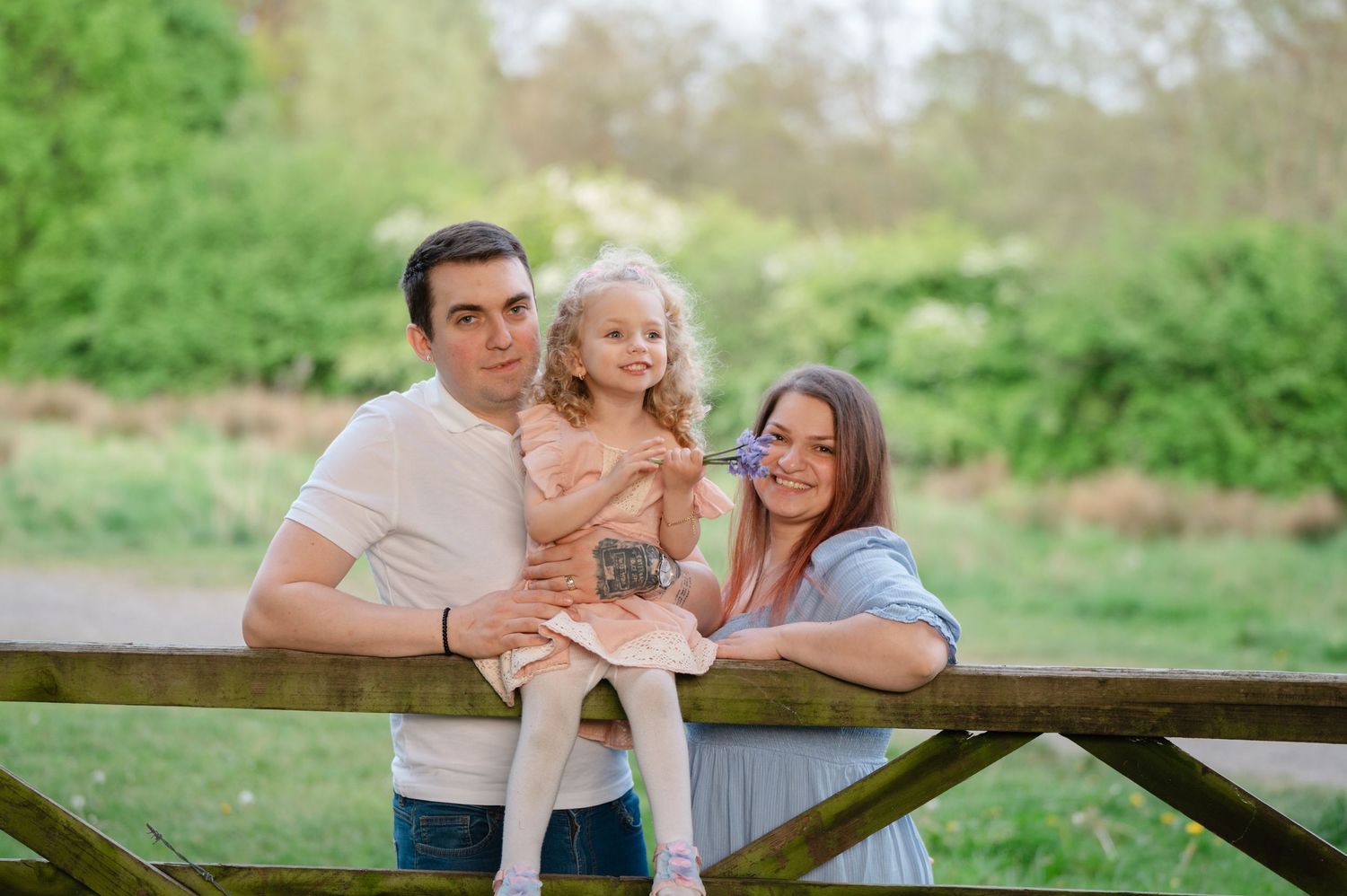 Family of three smiling together behind a rustic wooden fence in a meadow, with the young daughter in the center held by her parents.