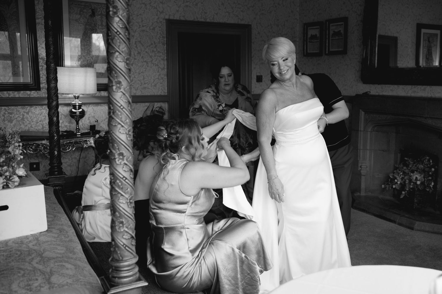 Black and white image of the bride surrounded by bridesmaids helping her into her wedding dress in a cosy, joyful pre-ceremony moment.
