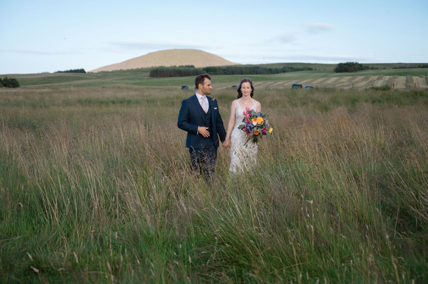 Bride and groom walking together through grassy meadow after ceremony