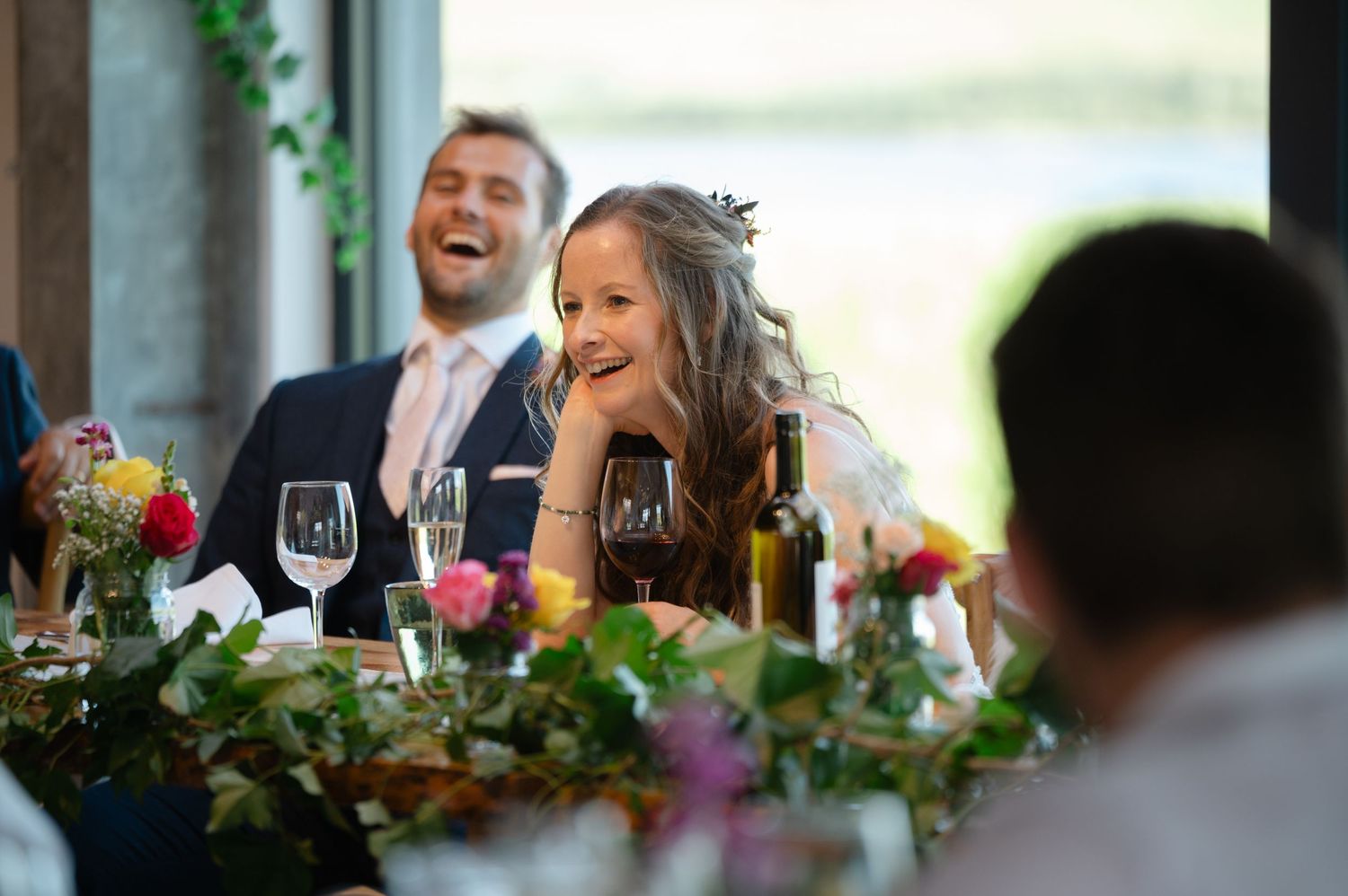 Bride laughing during wedding speeches surrounded by guests at reception table