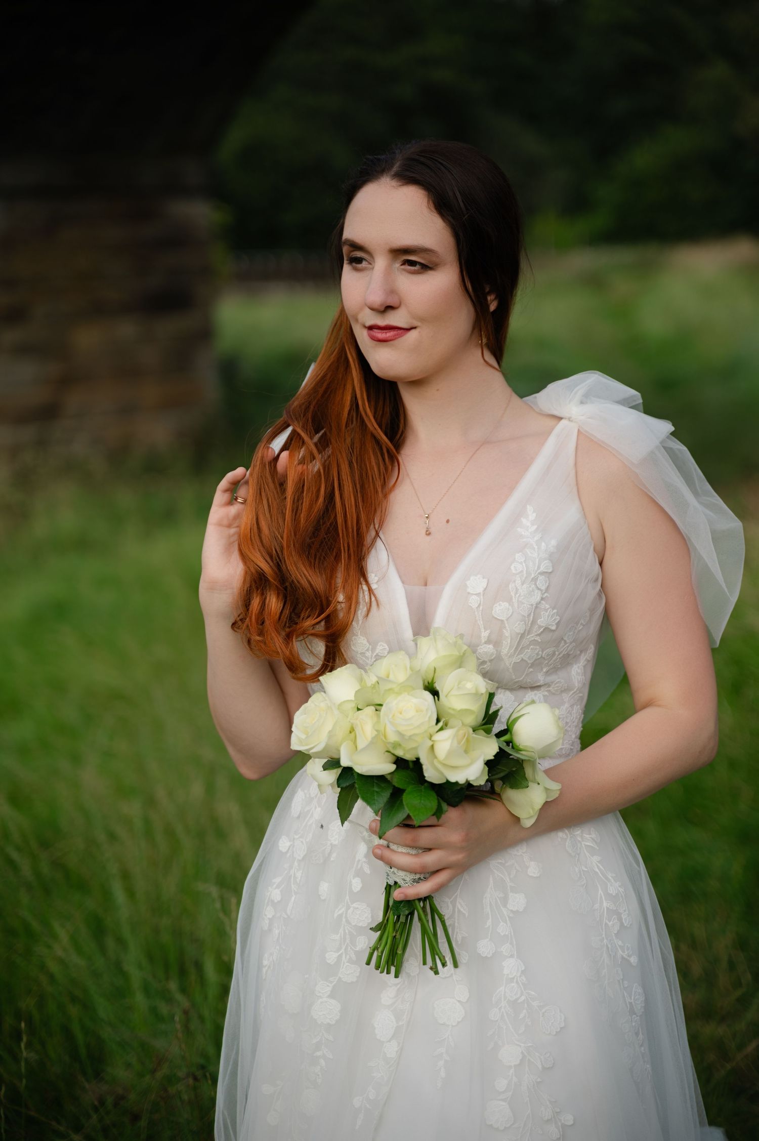 Bride with long red hair holding a white rose bouquet, smiling softly outdoors