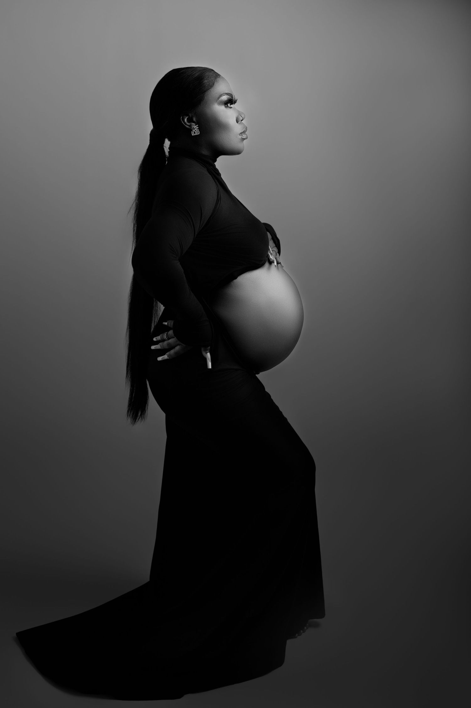 Side view of a pregnant woman in a long black dress, standing tall with hands under her belly against a dark backdrop.