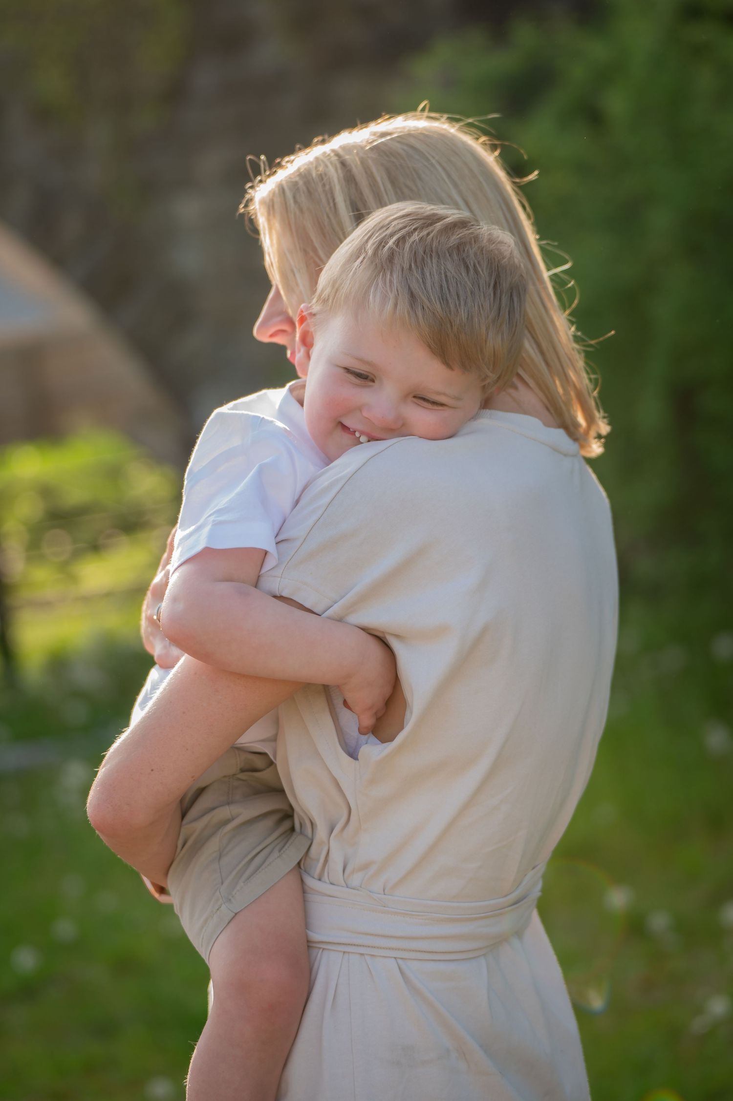 Older child gently hugging a younger sibling outdoors, both dressed in white and wrapped in a tender embrace with soft natural light.