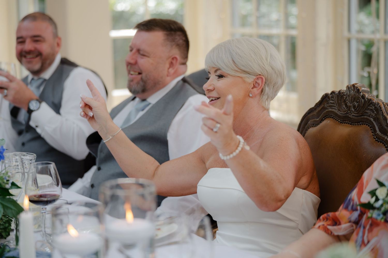 Bride and groom laughing with guests during speeches at the wedding head table, surrounded by drinks, florals, and joyful energy.