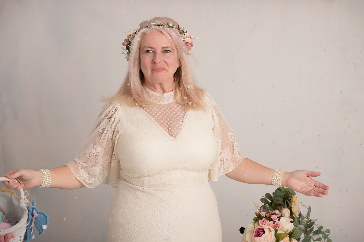 Bride in a lace wedding dress and floral crown, standing with arms slightly outstretched, smiling in a softly lit studio with floral decor.