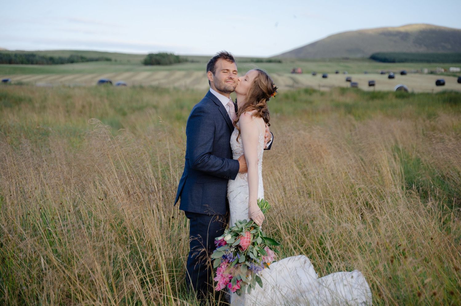Bride and groom kissing in tall grass with scenic hills in background