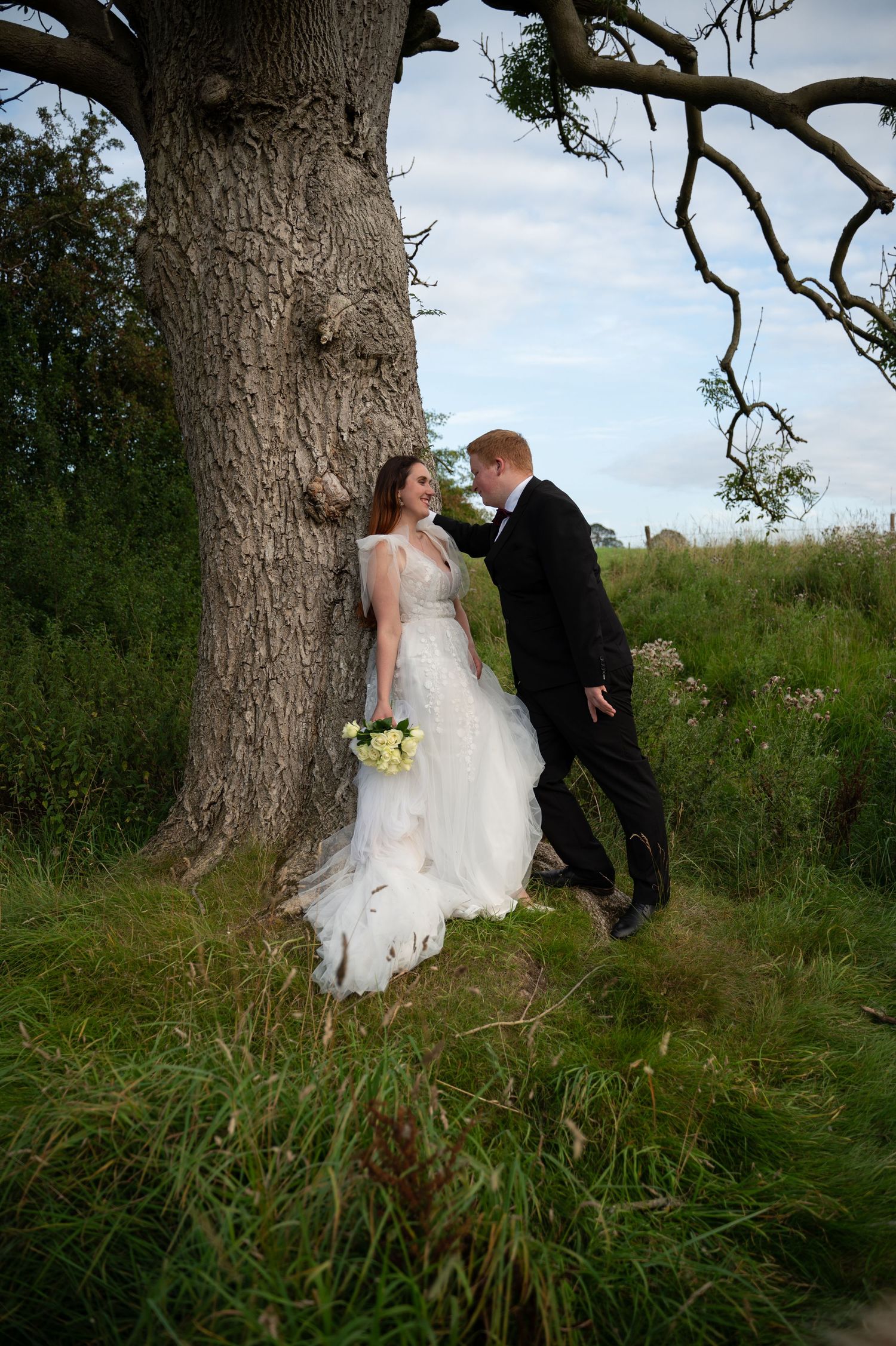 Bride and groom standing under a large tree, surrounded by tall grass