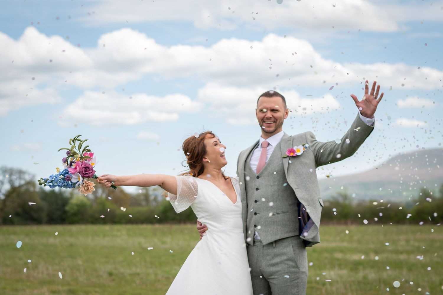 Bride and groom joyfully celebrating in an open field under a blue sky with scattered clouds, with mountains and trees in the distance.