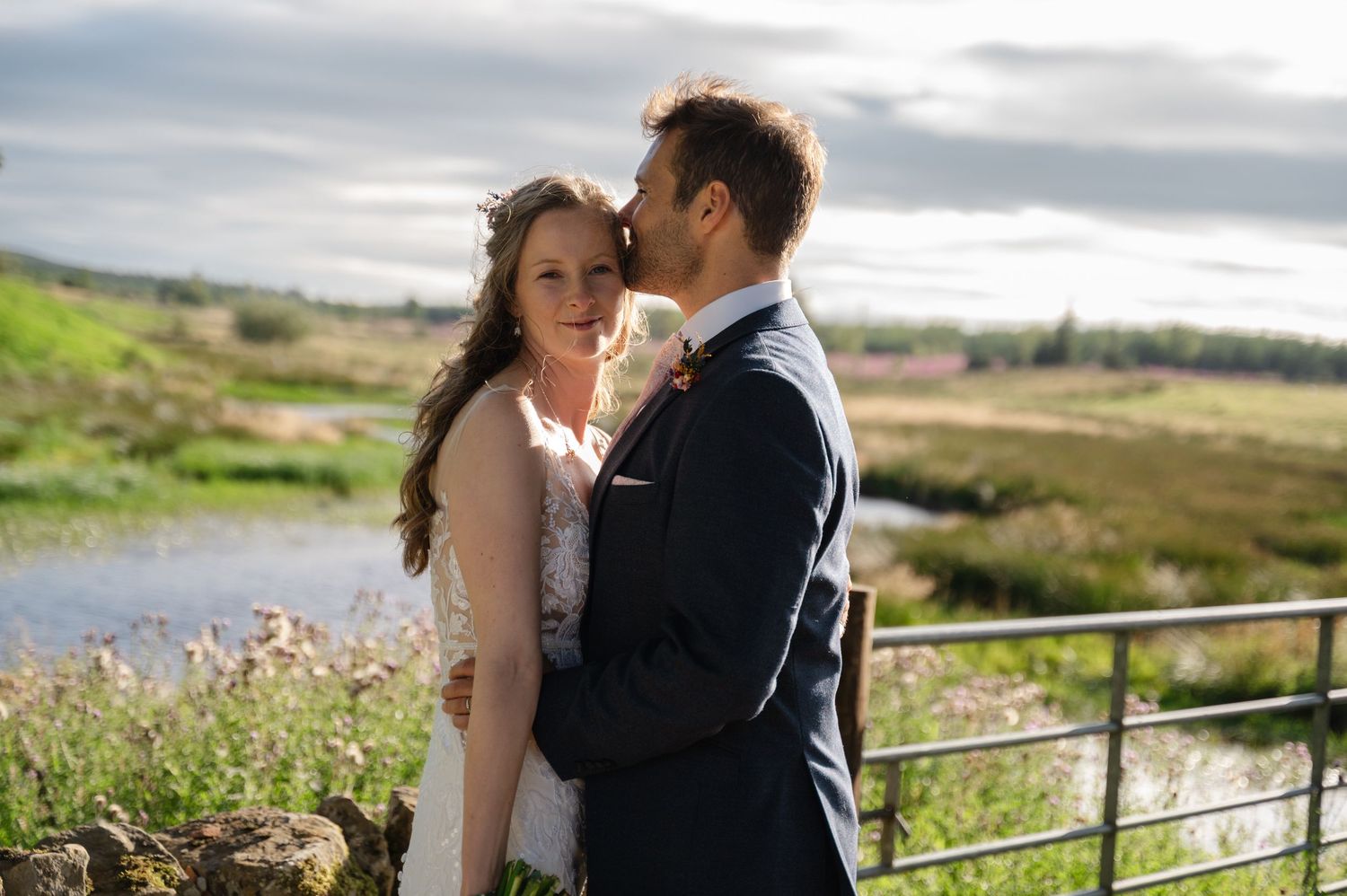 Groom kissing bride’s forehead as they embrace near waterside