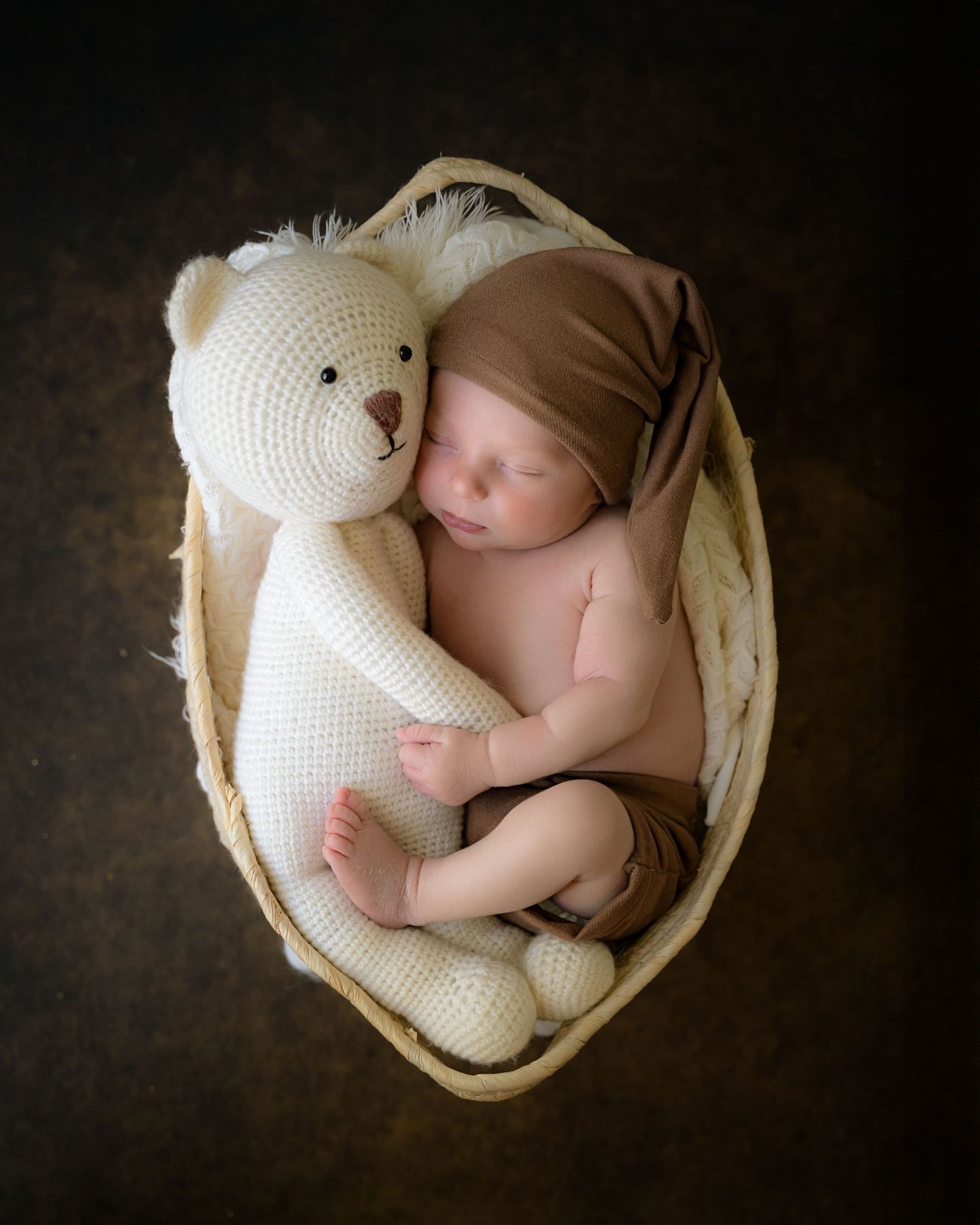 Newborn baby curled up in a small cream-colored basket with a white teddy bear, wrapped in a soft blanket.