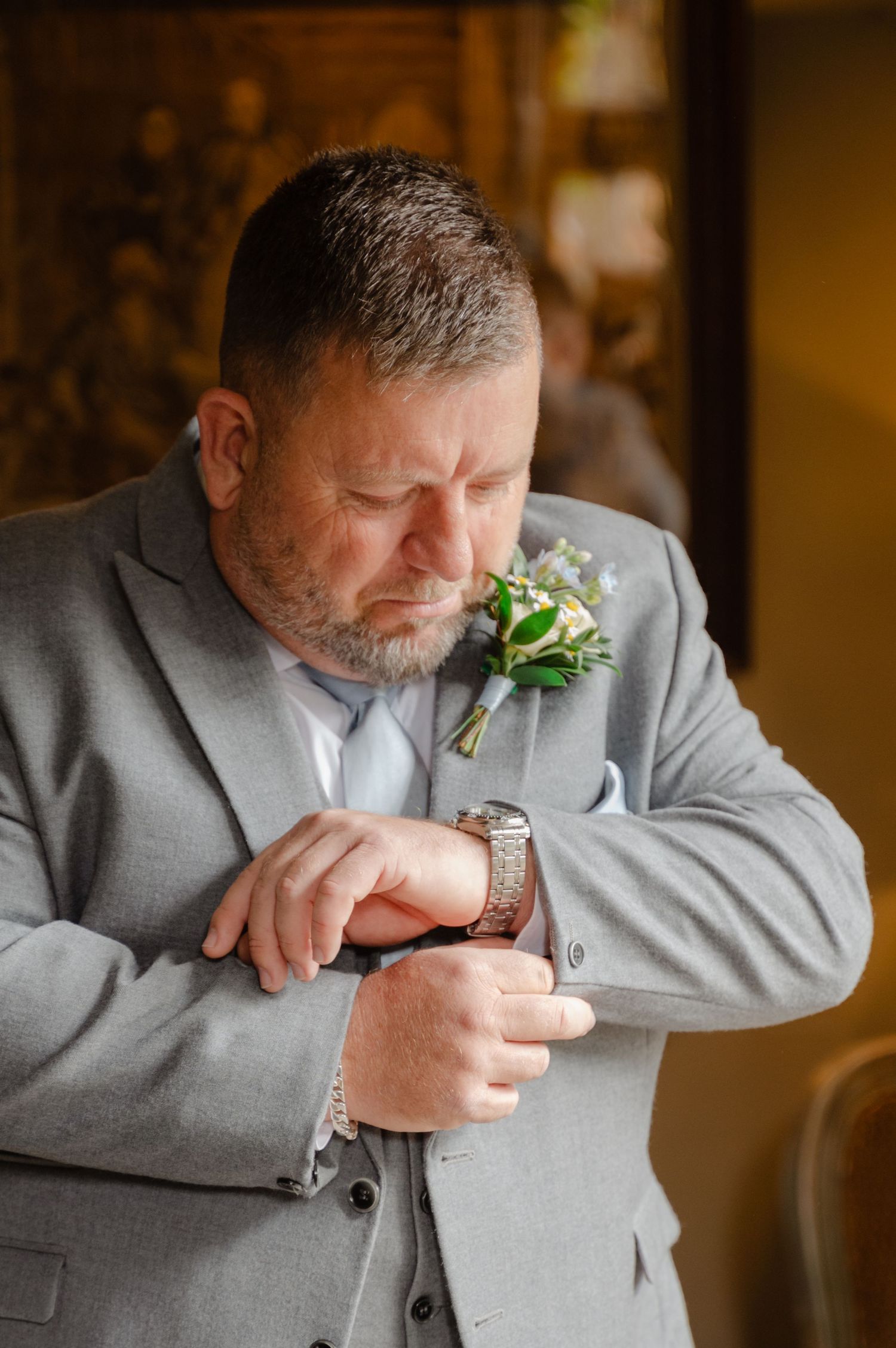 Groom carefully pinning a boutonnière onto his jacket, focused and preparing for the ceremony in a softly lit room.
