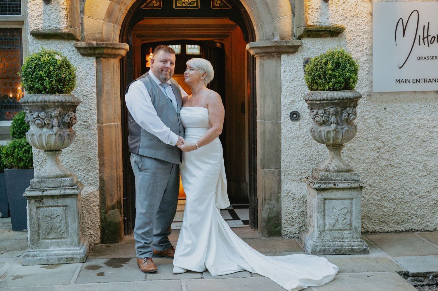 Bride and groom standing in front of an ornate stone archway at a wedding venue, holding each other and smiling in elegant attire.