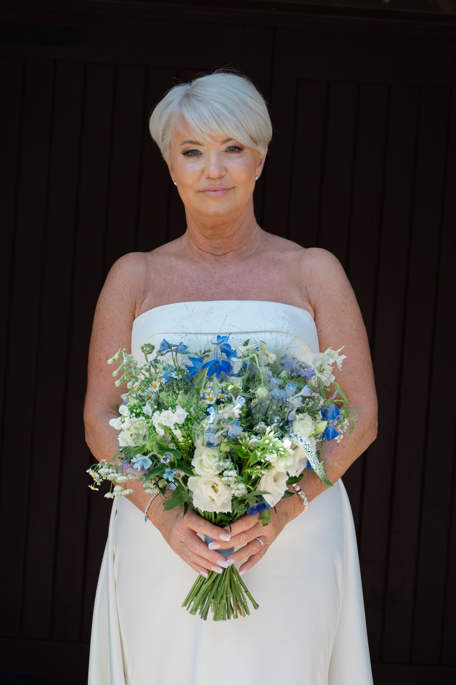Bride with short blonde hair holding a blue and white bouquet, standing against a dark background