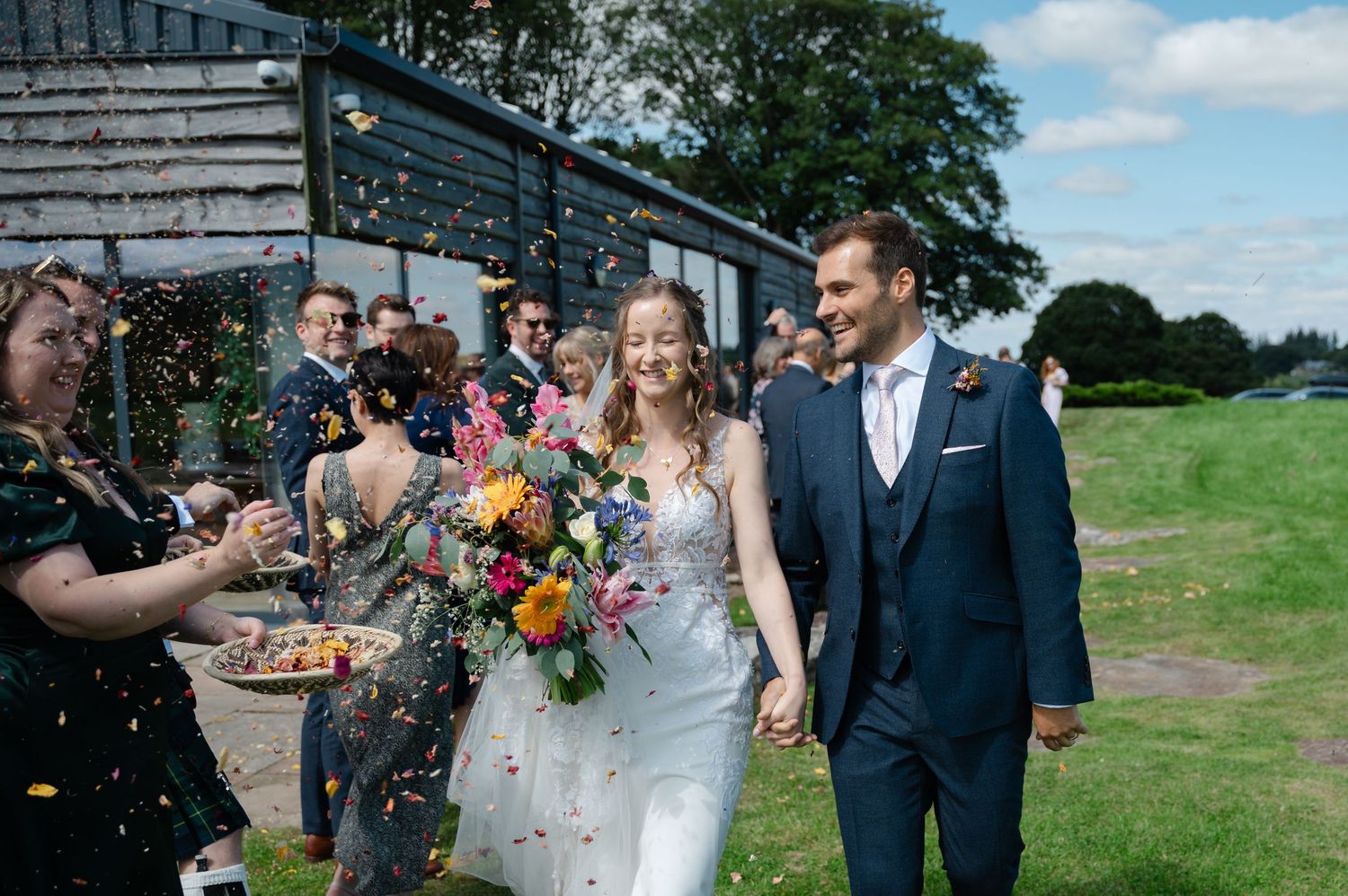Bride and groom walking as guests cheer and throw confetti outside rustic barn venue