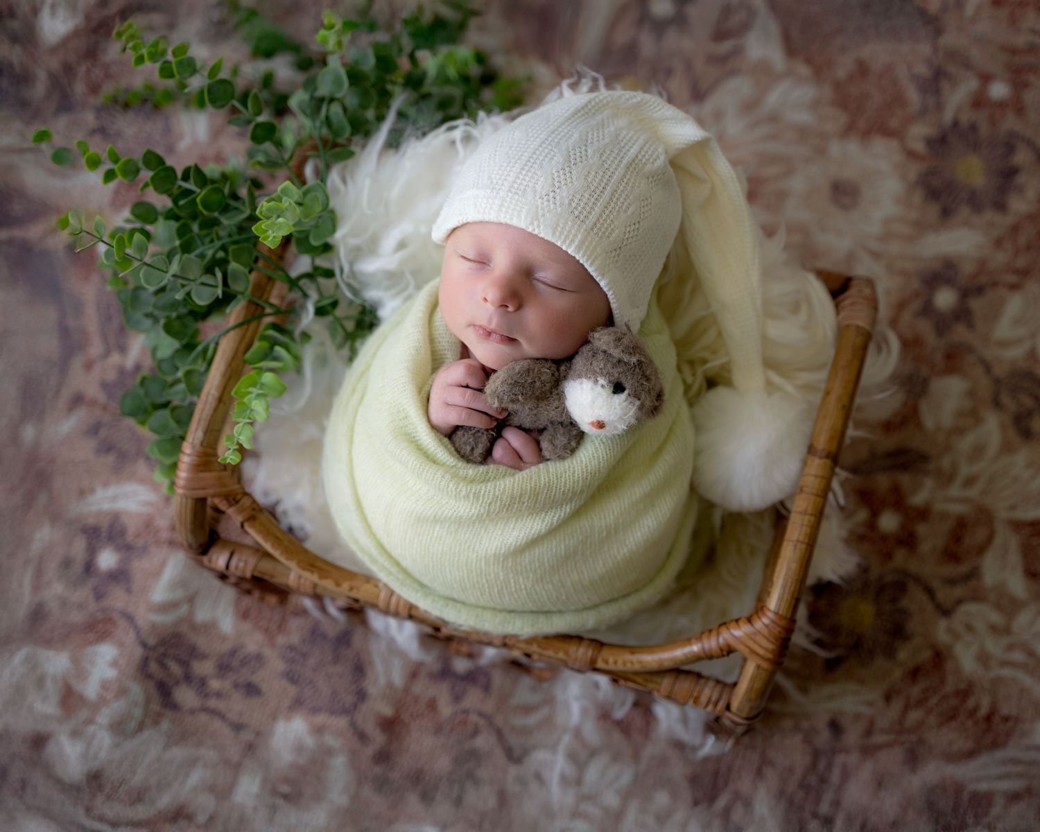 Newborn wrapped in a soft green blanket, lying on a miniature wooden bed with a tiny teddy bear, surrounded by greenery.