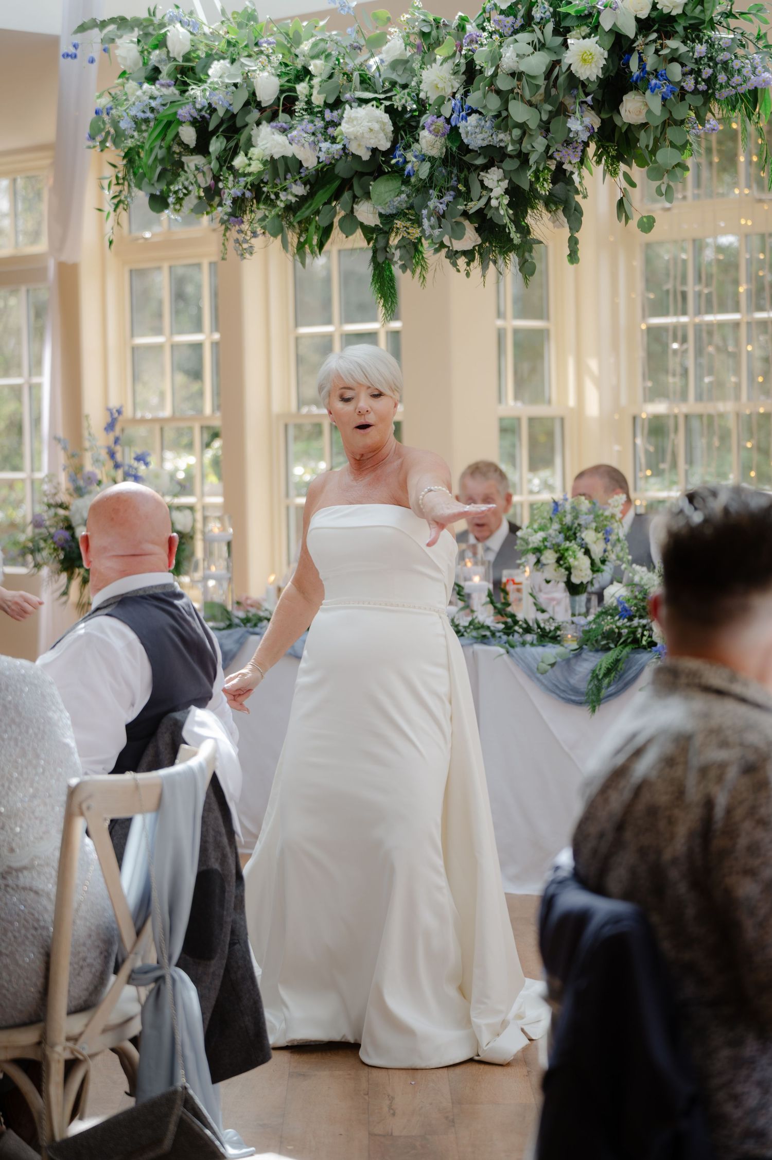 Bride spinning joyfully on the dance floor during the reception, with guests seated around and floral greenery hanging overhead.