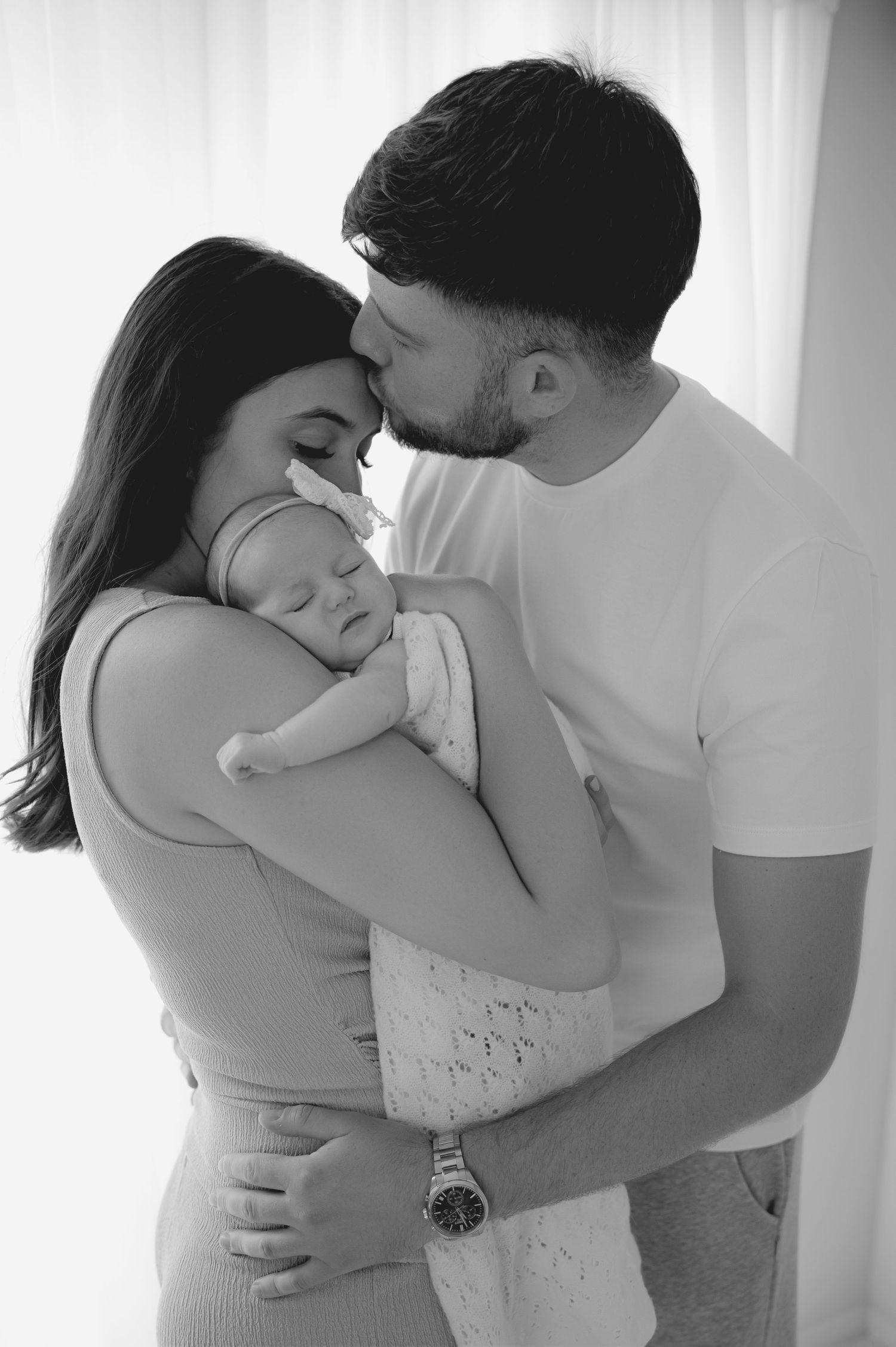Father gently kissing his newborn baby on the forehead while mother holds the baby, sharing a quiet, emotional family moment.