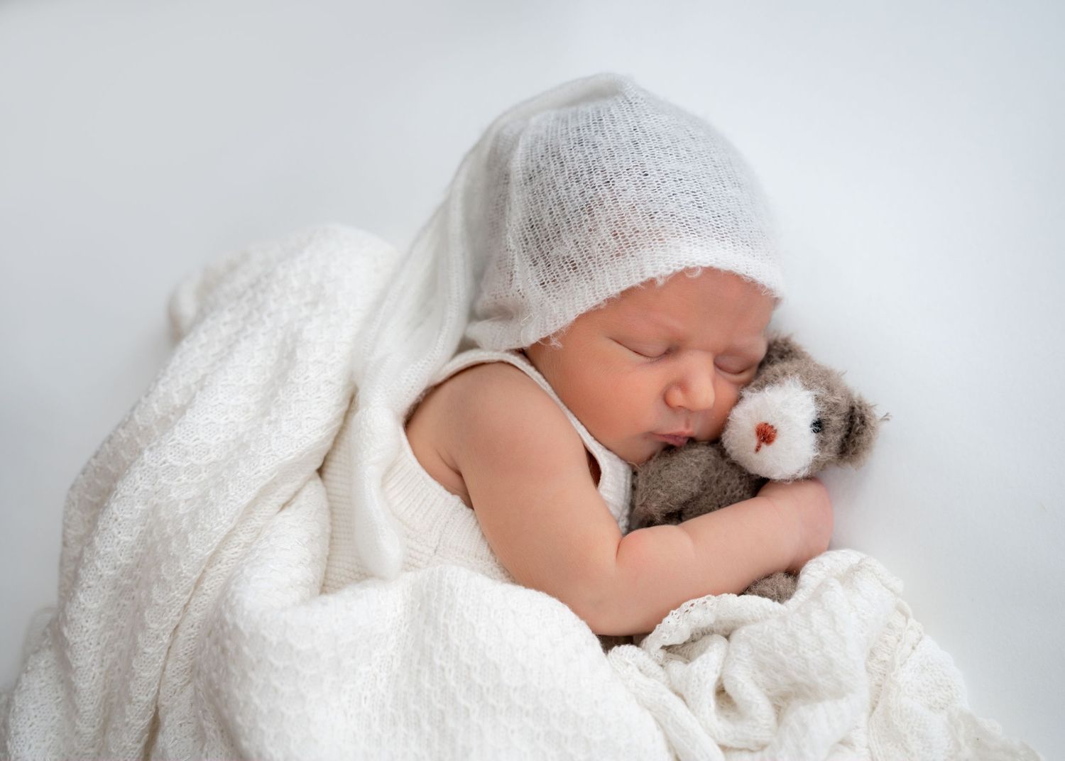 Newborn baby wrapped in white with a bonnet, holding a small teddy