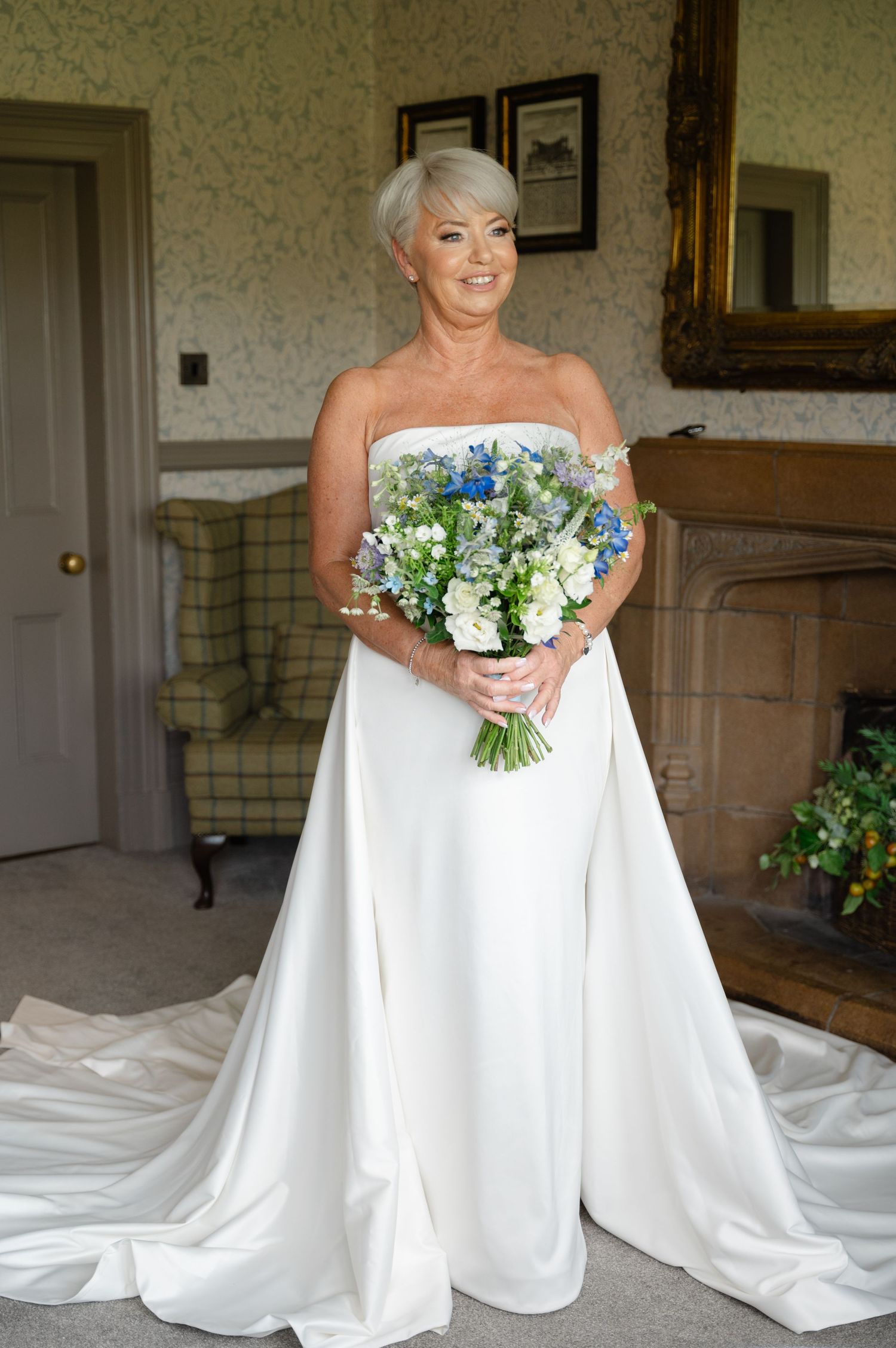 Bride posing in a full-length portrait, holding a bouquet of white and green flowers, standing in a warmly lit room before heading to the ce