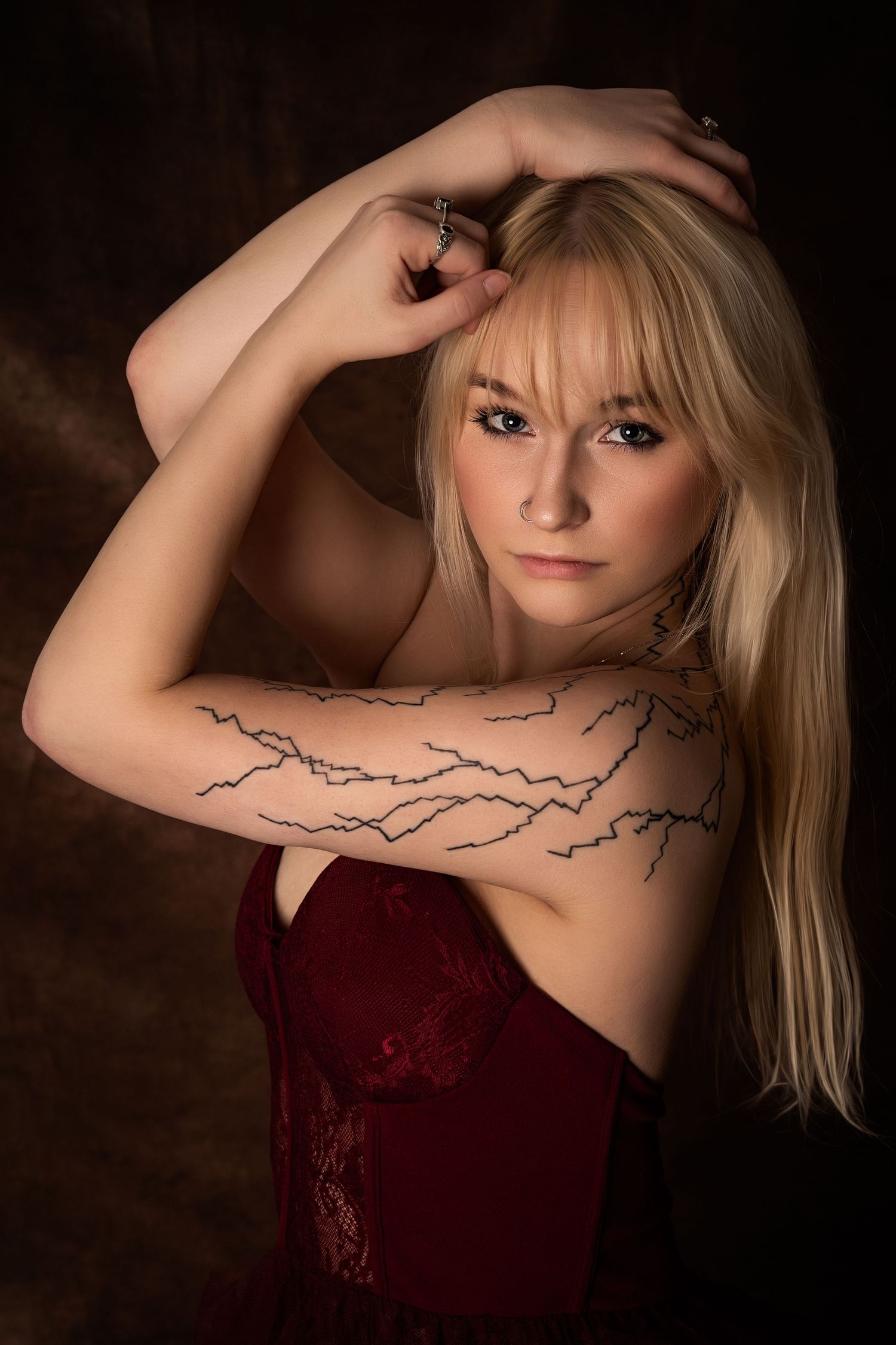 Studio portrait of a blonde woman in a strapless burgundy dress, posing with one arm raised, lit against a dark background with soft shadows