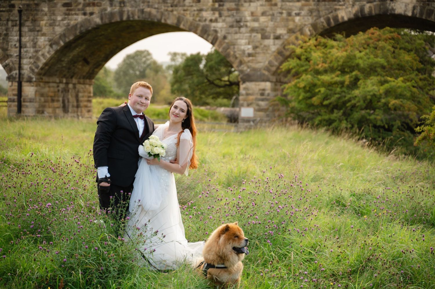 Bride and groom holding hands with their dog, standing in front of a stone viaduct